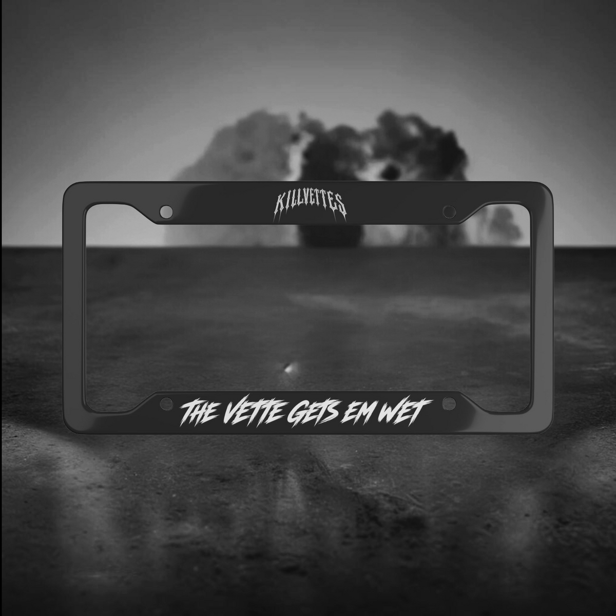 A black metal license plate frame with white stylized text reading 'KILLVETTES' at the top and 'THE VETTE GETS EM WET' at the bottom, displayed against a dark gray and black background with a blurred explosion behind it.